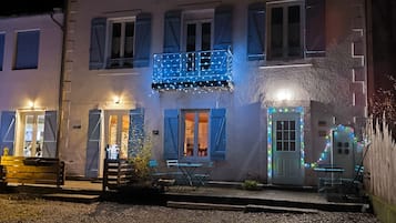 Een gebouw met twee verdiepingen met een balkon verlicht met lichtslingers, een deur met een klein portaaltje en een bankje buiten.