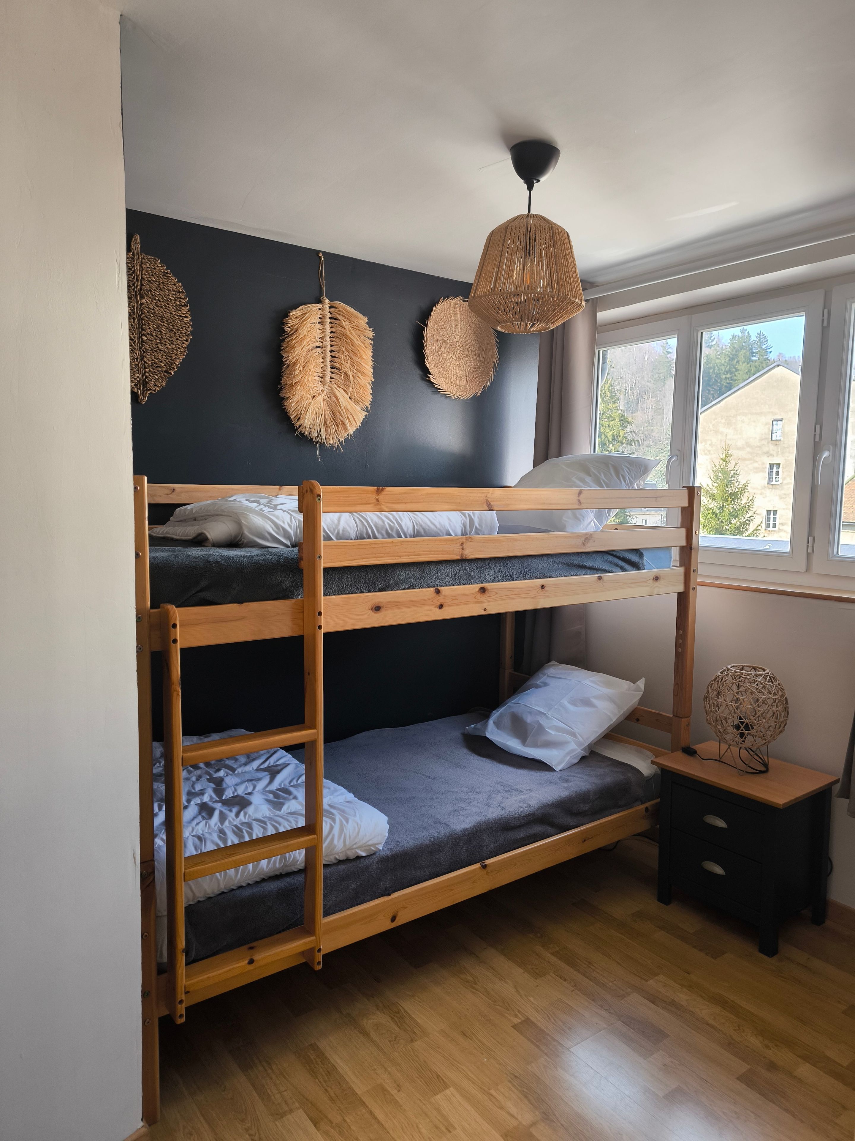 Een stapelbed met houten frame, een nachtkastje met lamp en een raam met uitzicht op gebouwen.