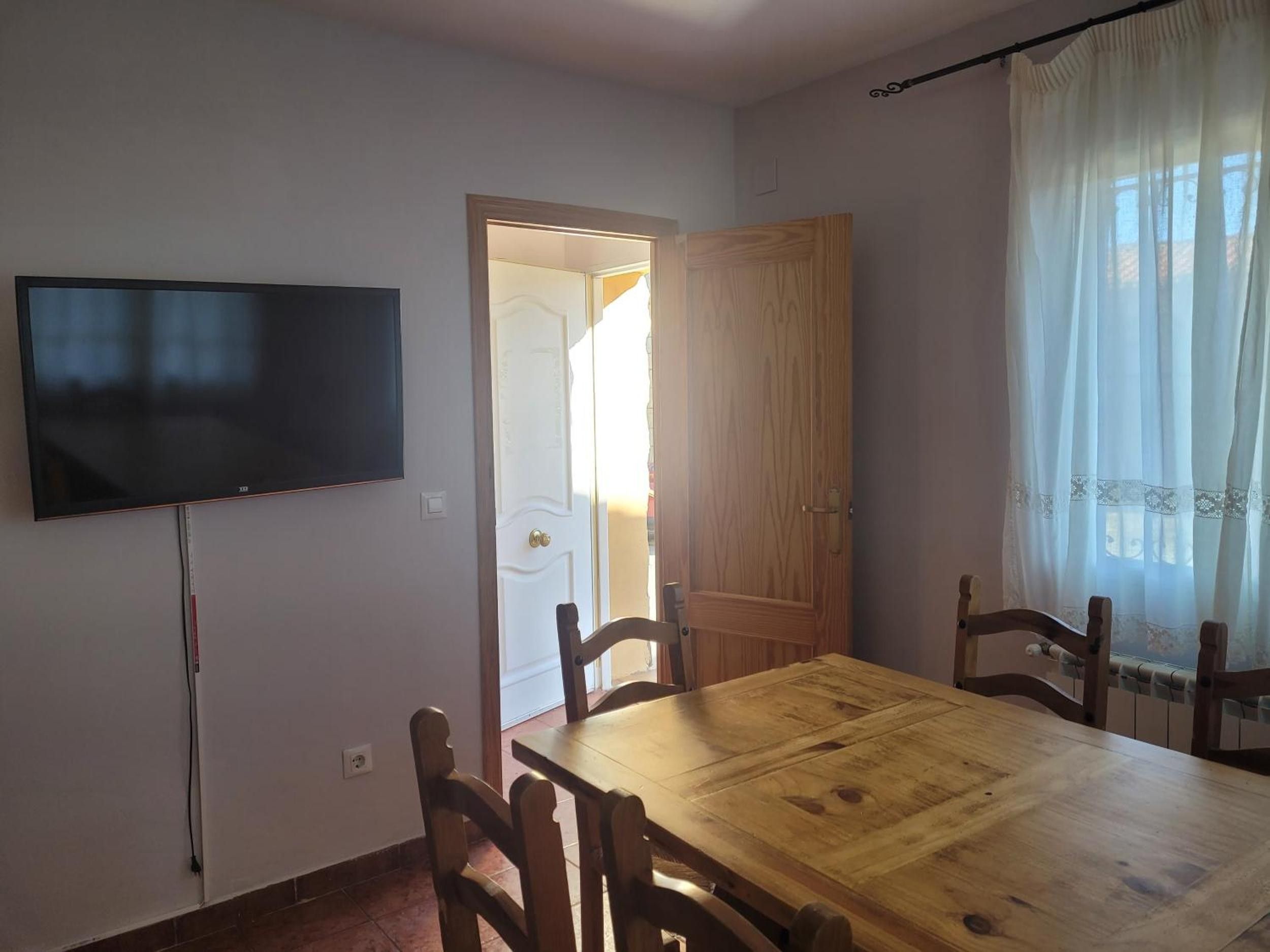 Een eetgedeelte met een houten tafel en stoelen, een aan de muur bevestigde televisie en een deur naar een andere kamer.