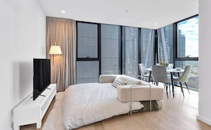 Une chambre moderne dotée d’un grand lit, d’une télévision, d’un coin repas avec une table et des chaises, et offrant une vue sur la ville grâce à de grandes fenêtres.