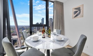 Un coin repas avec une table dressée pour deux personnes, une bouteille de spiritueux et une vue sur le paysage urbain à travers de grandes fenêtres.