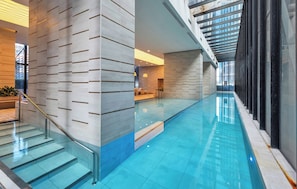 Une piscine intérieure moderne, dotée d’une paroi en verre, d’un escalier avec une rampe métallique et d’un espace d’accueil avec un canapé et une plante.