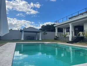 Una piscina con un porche cubierto, un muro de piedra y un cielo azul despejado.