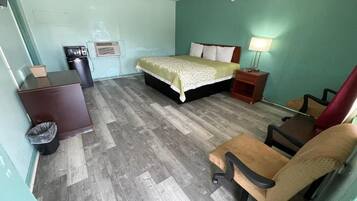 A room with a bed, a desk, a chair, a trash bin, and a nightstand with a lamp.