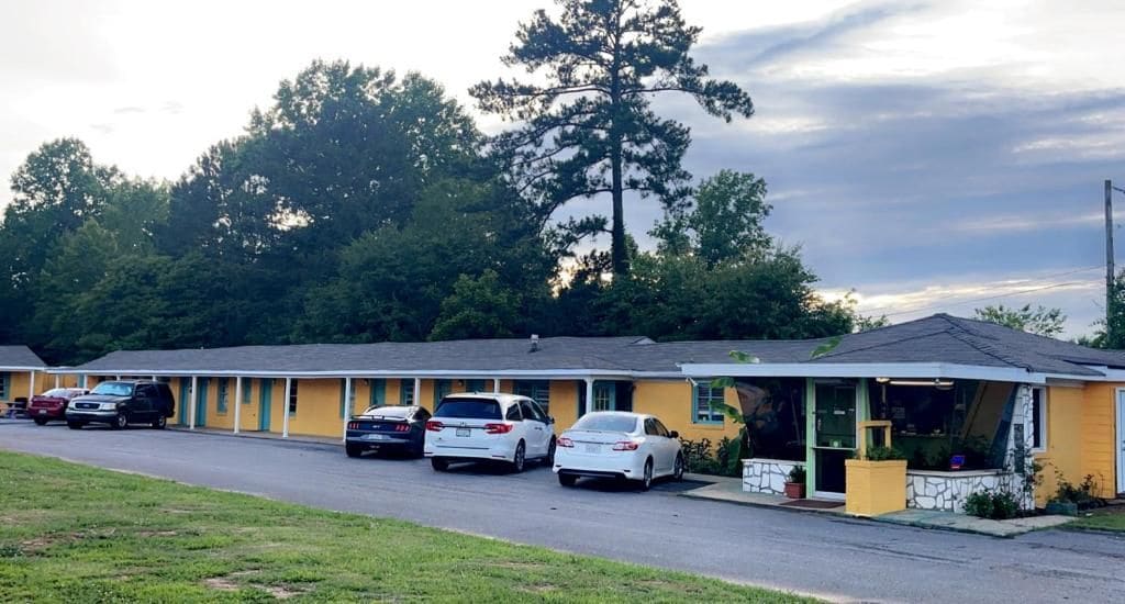 A motel with a row of parked cars, a single-story building, and a large tree in the background.