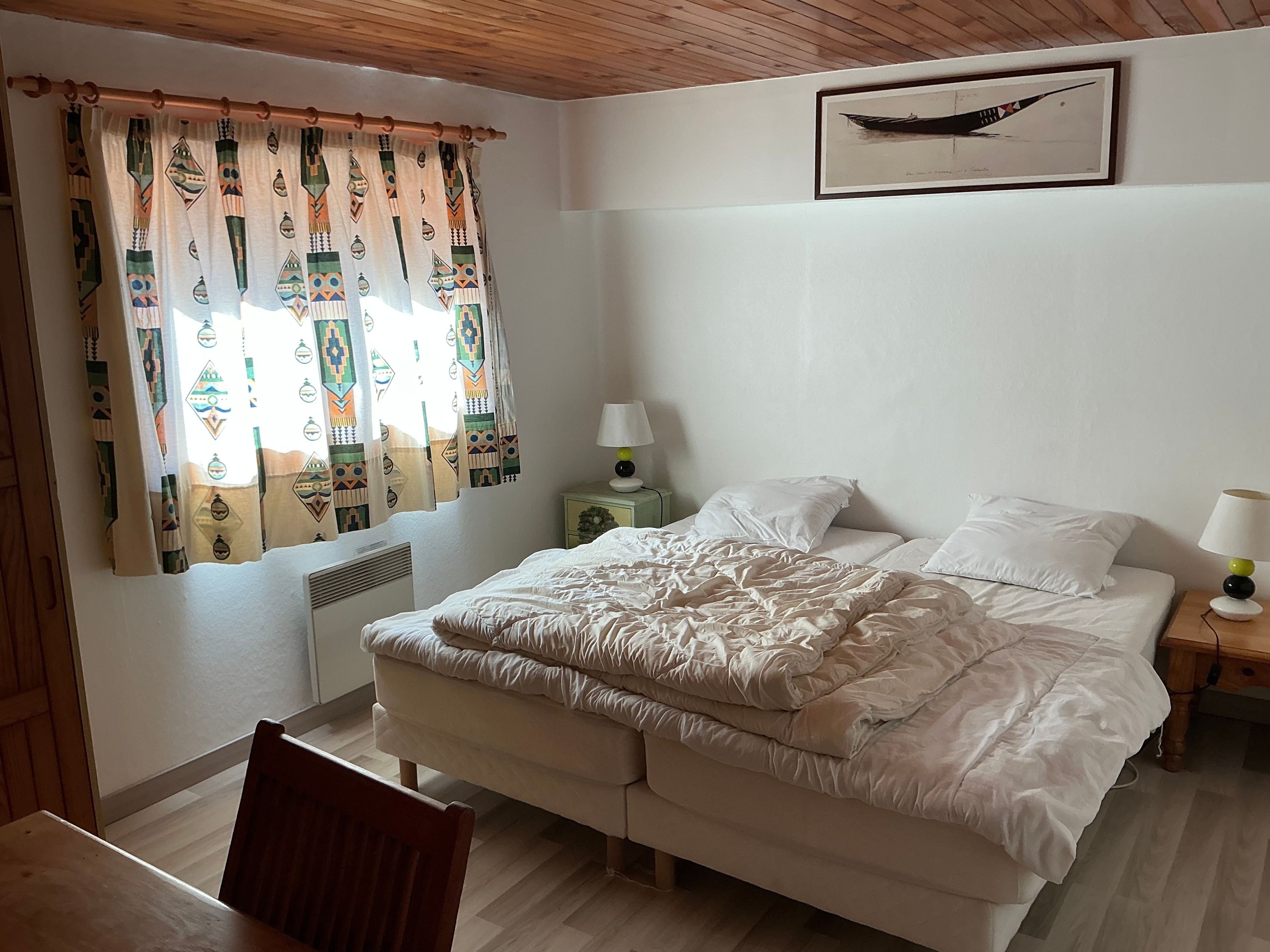 Une chambre à coucher avec un lit, des tables de chevet, une chaise, une fenêtre avec des rideaux et un tableau encadré au mur.
