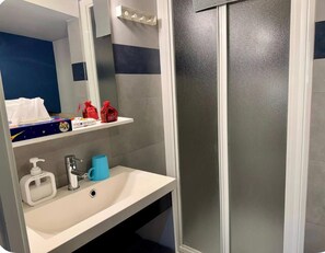 Une salle de bain avec un lavabo blanc, un miroir, un distributeur de savon bleu et une douche dotée d’une porte en verre dépoli.
