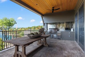 An outdoor patio with a wooden table and chairs, a grill, and a view of a body of water. - Crimson Waters at Hill House | Golf Cart Optional (Tuscaloosa)
