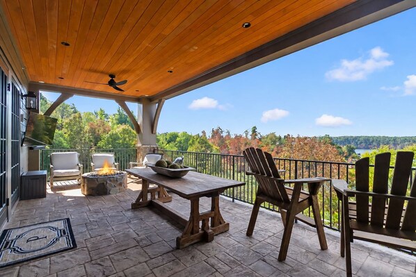 An outdoor patio with a wooden ceiling, stone flooring, and a view of a lake surrounded by trees. - Crimson Waters at Hill House | Golf Cart Optional (Tuscaloosa)