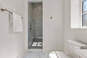 A bathroom with a glass shower enclosure, white marble tiles, a white toilet, and a window. - The Retreat at Hill House (Tuscaloosa)