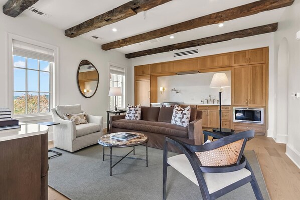 A modern living room with a sofa, armchair, and coffee table, a kitchen with wooden cabinets, and a large window. - The Retreat at Hill House (Tuscaloosa)