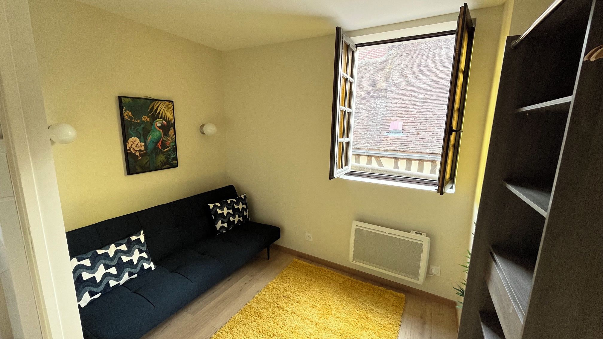 A small living room with a black sofa, a yellow rug, a radiator, and a window with a view of a brick building.