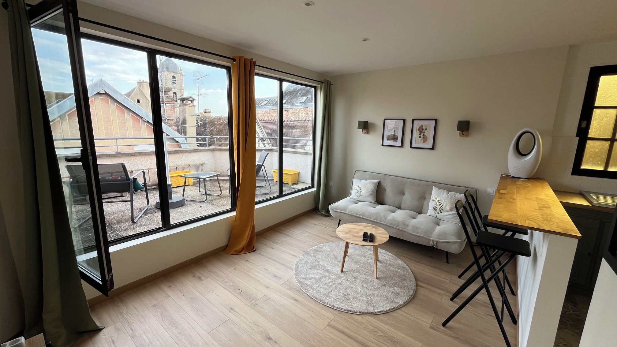 A modern living room with a large window, a grey sofa, a round coffee table, and a dining area with a wooden table and chairs.