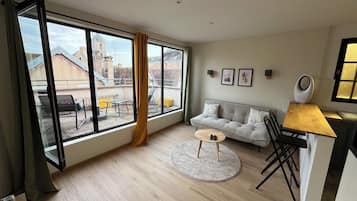 A modern living room with a large window, a grey sofa, a round coffee table, and a dining area with a wooden table and chairs.
