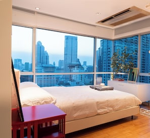 Un dormitorio con una cama grande, un escritorio y vistas a un paisaje urbano a través de ventanas de piso a techo.