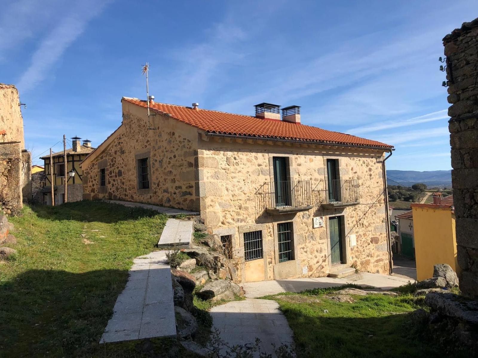 Un bâtiment en pierre avec un toit recouvert de tuiles rouges, un petit balcon et un chemin menant à l’entrée.