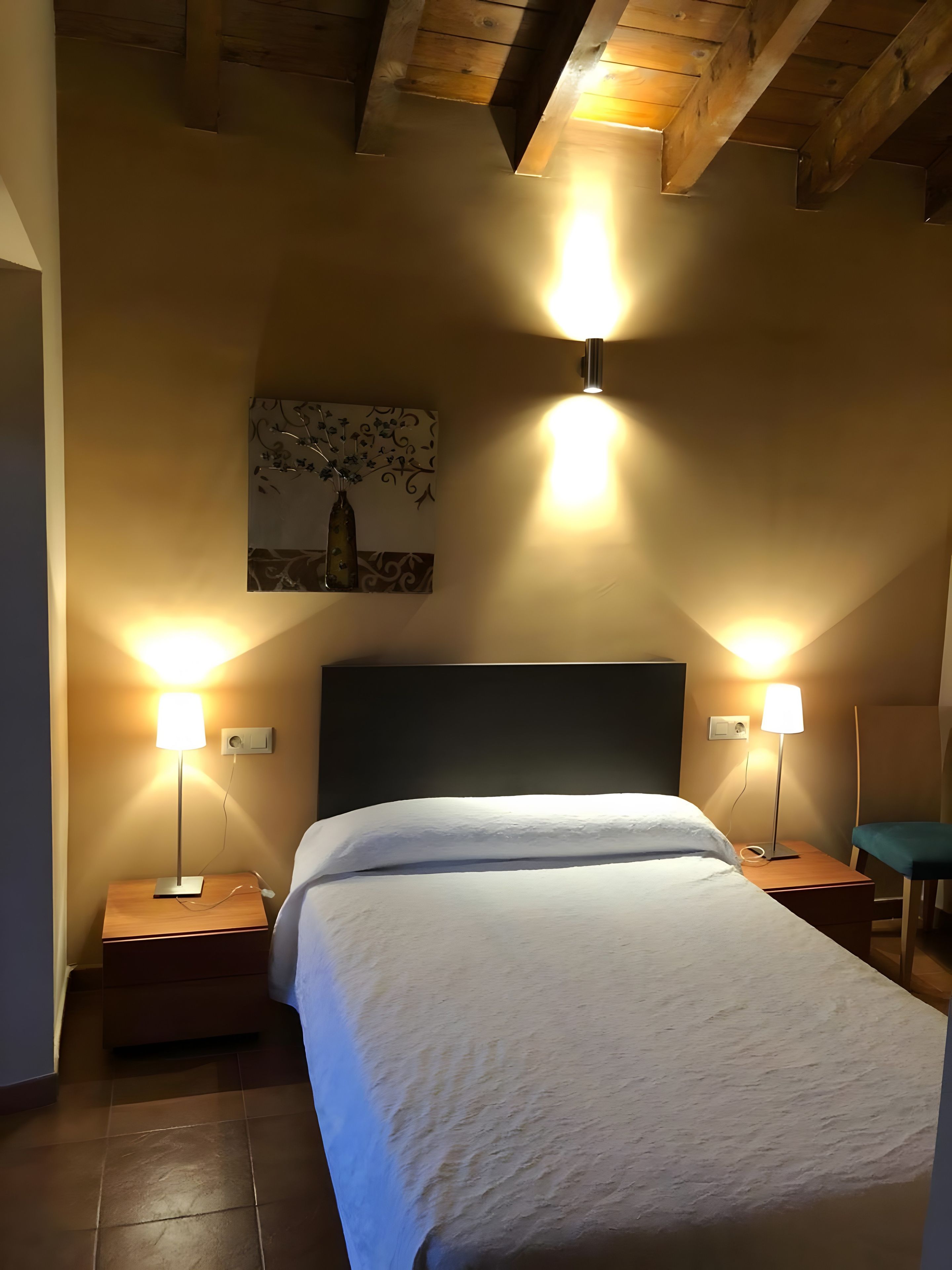 Une chambre à coucher avec un lit, deux tables de chevet, deux lampes fixées au mur, un tableau accroché au mur et un plafond en bois.
