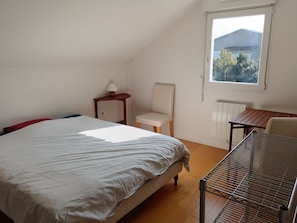 Une chambre à coucher comprenant un lit, une chaise, une petite table, un radiateur et une fenêtre donnant sur des immeubles et des arbres.