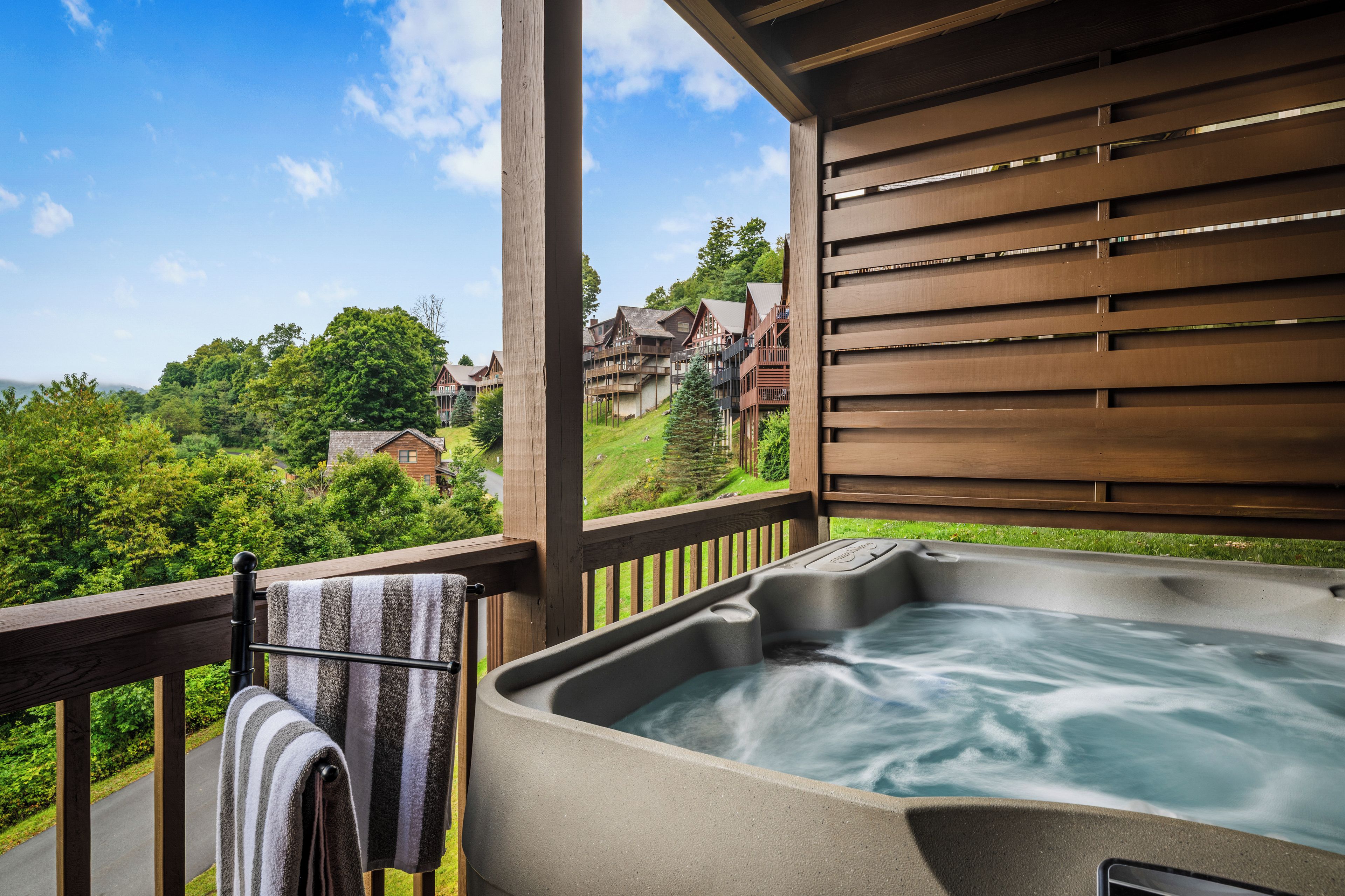 Un jacuzzi en una terraza de madera con una toalla colgada en un perchero, con vistas a una zona boscosa y edificios.