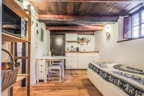 Une pièce de taille modeste avec un plafond en bois, un lit recouvert d’une literie à motifs, une petite table avec une chaise, un coin cuisine avec des placards et une porte.