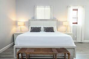 Un dormitorio ordenado con una cama grande, dos mesitas de noche con lámparas y un pequeño banco de madera frente a la cama.