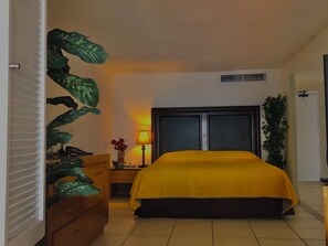 A hotel room with a bed, a nightstand, a lamp, a dresser, and a potted plant. - New Listing (Puerto Vallarta)