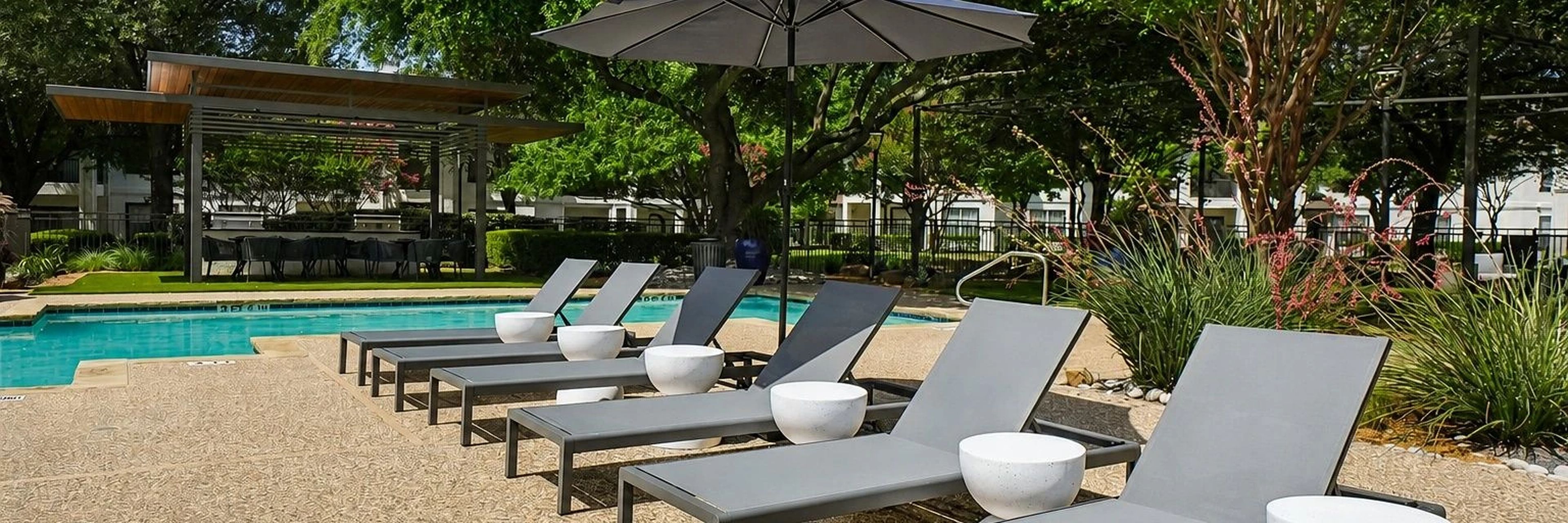 Area piscina con lettini e ombrelloni, circondata da vegetazione e un edificio sullo sfondo.