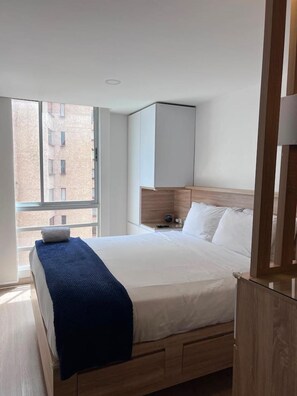 A hotel room with a bed, a nightstand, a wooden headboard, and a window with a view of buildings. - Premium Loft La Candelaria (Bogotá)