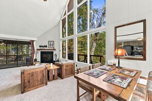 A spacious room with a large dining table, a fireplace, and large windows offering a view of trees.