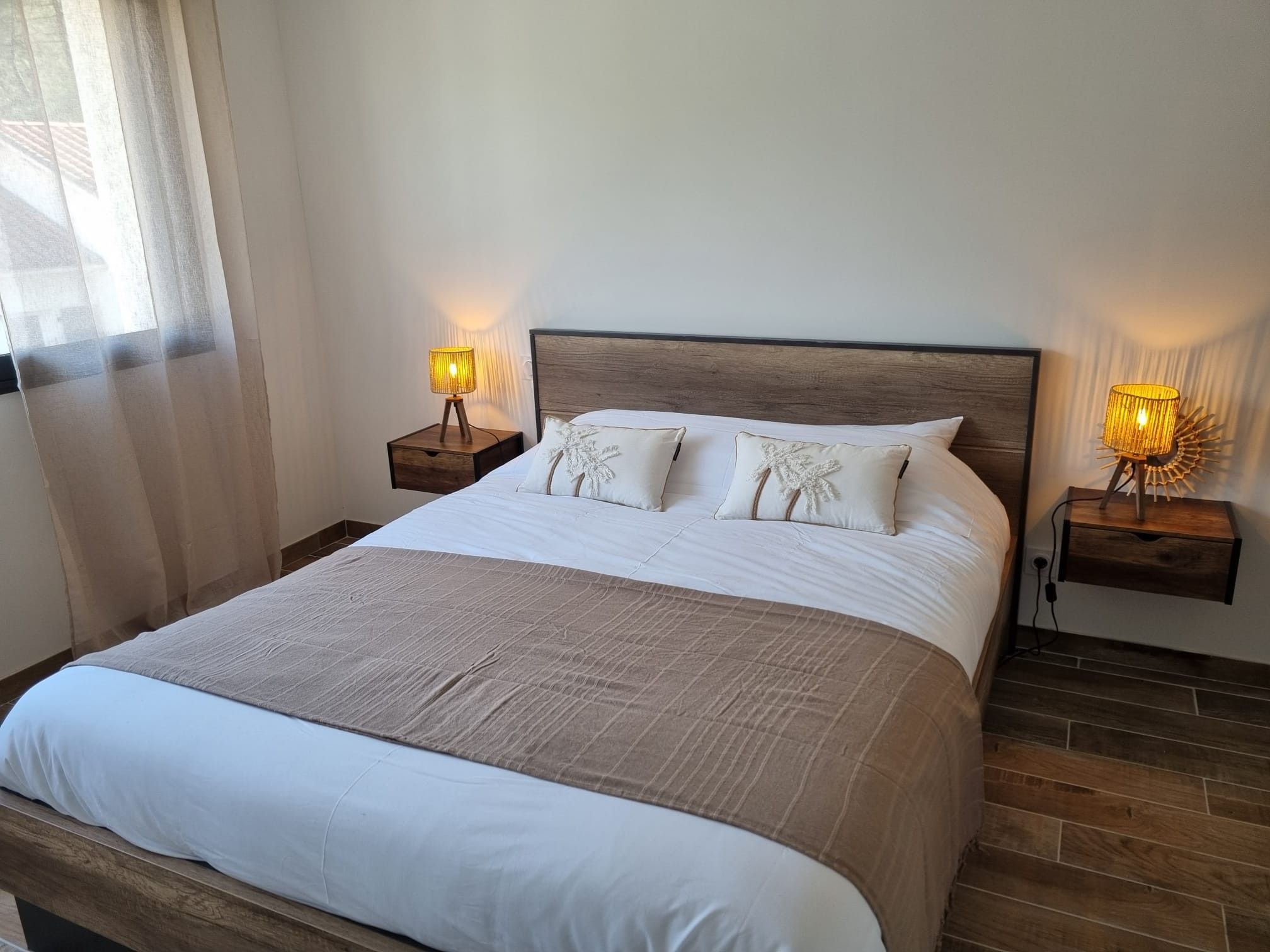A neatly made bed with a wooden headboard, two bedside tables with lamps, and a window with sheer curtains.