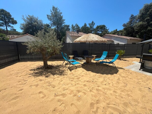 Un jardin avec un espace sablonneux, un arbre et des chaises longues bleues.