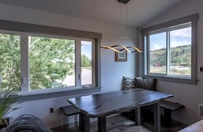 Un comedor moderno con una mesa larga y bancos, ventanas amplias que ofrecen vistas al verdor, y una lámpara contemporánea.