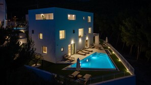 Un bâtiment moderne à deux étages, doté d’une piscine, d’un espace extérieur aménagé et de nombreuses fenêtres illuminées.