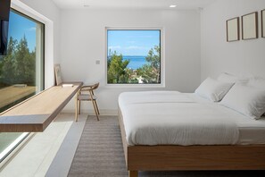 Une chambre à coucher avec un lit, un bureau, une chaise, une fenêtre donnant sur la mer et un tableau encadré au mur.