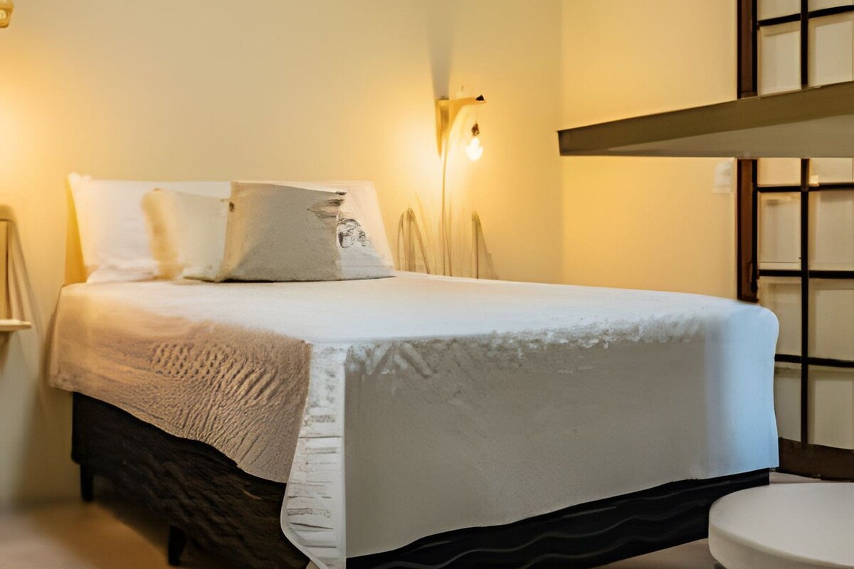 Una cama bien hecha con cubrecama y almohadas blancas, una lámpara de noche y una mesita redonda.