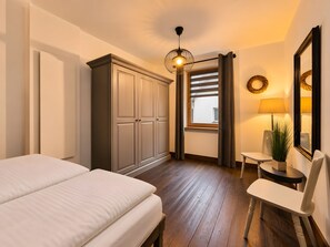 Ein Schlafzimmer mit Bett, Kleiderschrank, Nachttisch, Lampe, Stuhl und einem Fenster mit Vorhängen.
