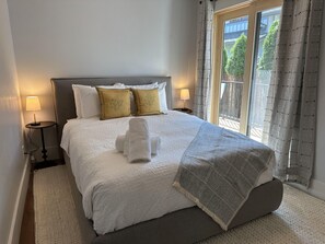 Een netjes opgemaakt bed met witte lakens en een grijze deken, twee nachtlampjes, en een raam met gordijnen.