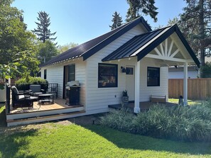 Een klein, gelijkvloers huis met een overdekte patio, een barbecue en een houten terras.
