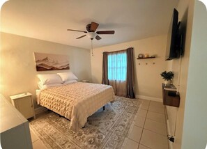 Ein Schlafzimmer mit einem Bett, einem Deckenventilator, einem Fenster mit Vorhängen, einem Fernseher und einer kleinen Ablagefläche mit Gegenständen.
