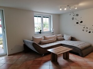 Ein modernes Wohnzimmer mit einem Ecksofa, einem kleinen Holztisch und einem Fenster mit Blick auf Grünflächen.