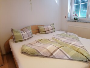 Ein ordentlich bezogenes Bett mit einer Karo-Decke und zwei Kissen, ein Holzkopfschild, ein Fenster mit Jalousien und eine kleine Pflanze auf der Fensterbank.
