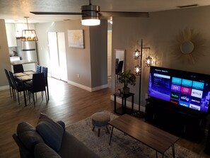 Una sala de estar moderna con un televisor de pantalla plana mostrando la interfaz de un servicio de streaming, un área de comedor con una cocina al fondo y un ventilador de techo.