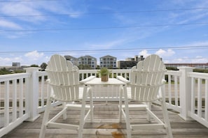 Une terrasse en bois avec deux chaises Adirondack blanches et une petite table, donnant sur un quartier résidentiel avec des maisons et un ciel dégagé.