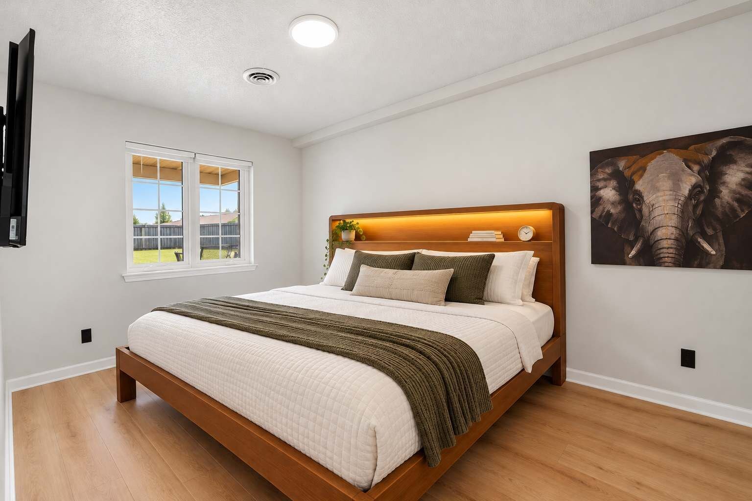 A bedroom with a bed, a window, a wall-mounted TV, and a painting.