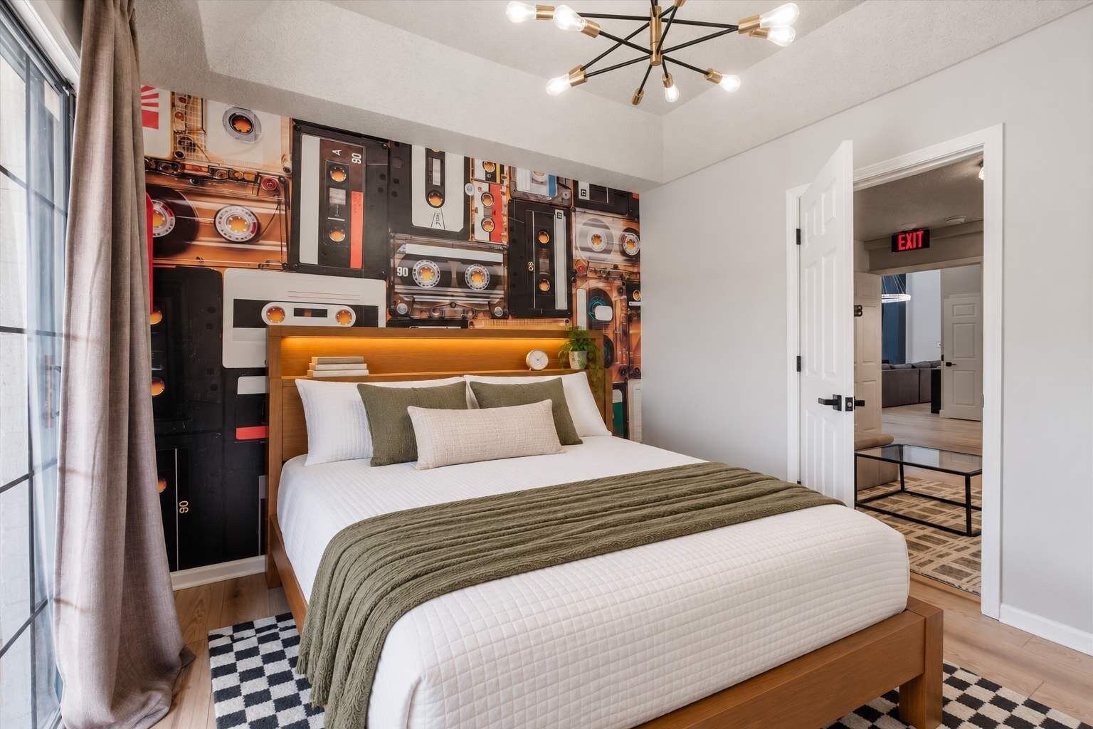 A modern bedroom with a bed, a headboard with a clock, a wall with cassette tapes, and an exit sign.