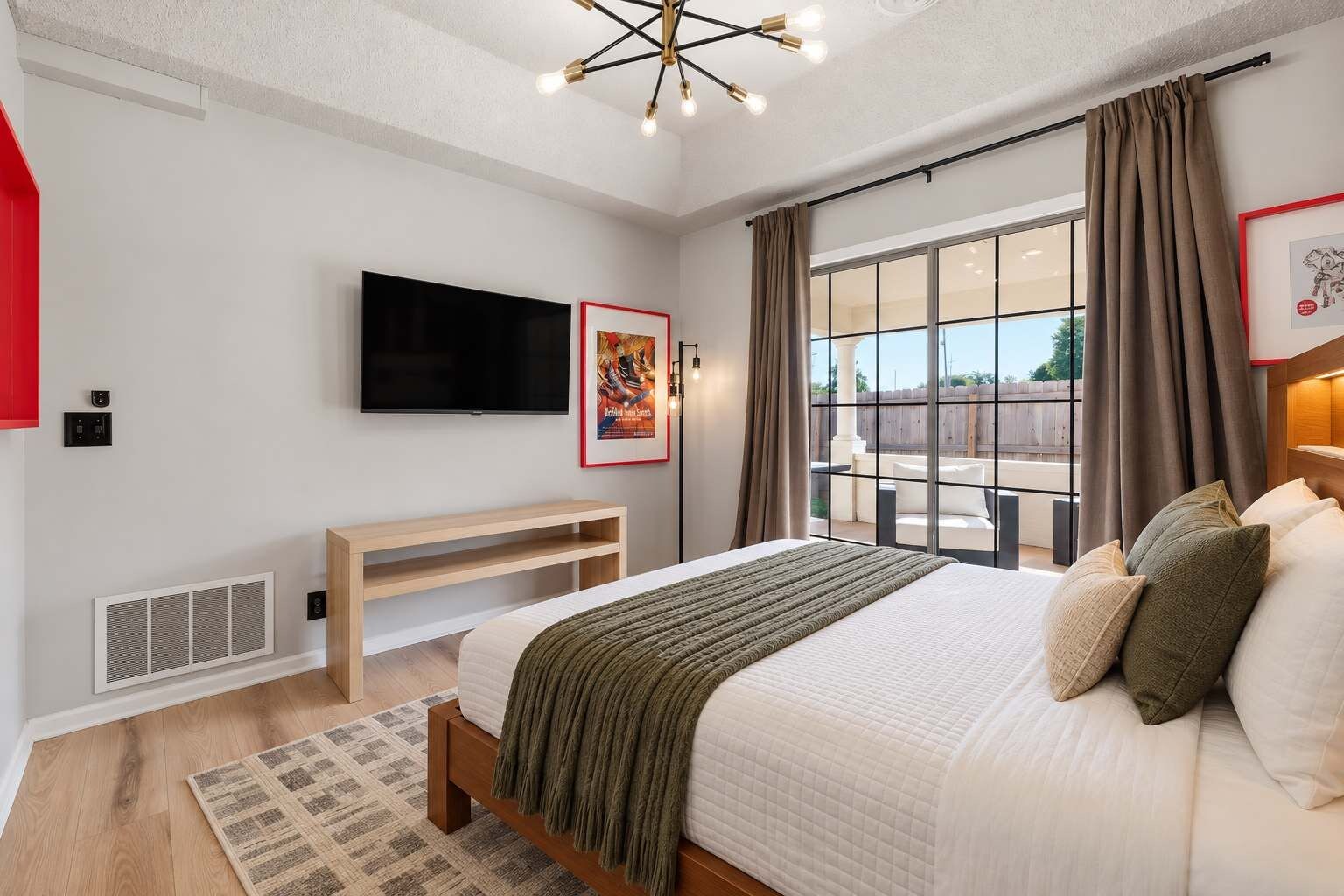 A modern bedroom with a large bed, a flat-screen TV, a wooden bench, and a view of the outdoors through a large window.