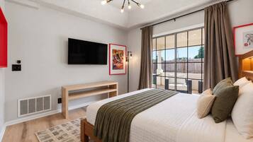 A modern bedroom with a large bed, a flat-screen TV, a wooden bench, and a view of the outdoors through a large window.