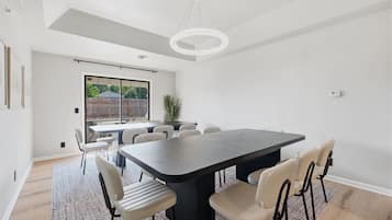 A modern dining area with a large table and chairs, a chandelier, and a view through a large window.
