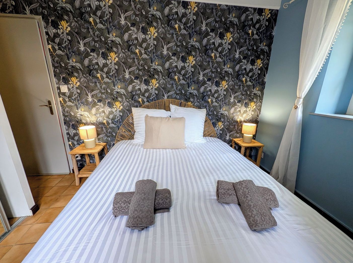 Une chambre d’hôtel avec un lit, deux tables de chevet, une porte et un papier peint à motifs floraux.