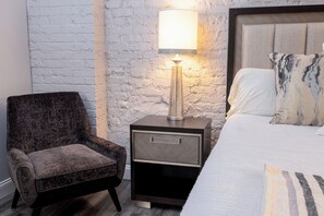 A bedroom with a bed, a nightstand with a lamp, a chair, and a brick wall. - Cozy 1-bedroom hotel suite in beautiful Seneca with AC - ROOM 4 (Seneca)